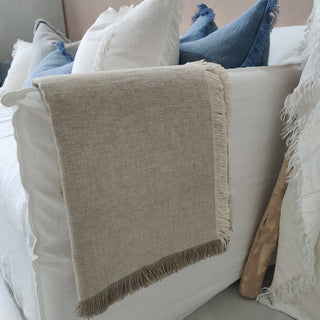 Beige throw blanket with fringe draped over a white sofa with blue and gray pillows.